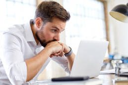 Mann in weißem Hemd arbeitet nachdenklich am Laptop an Schreibtisch, Schreibtischlampe leuchtet, Bürohintergrund sichtbar.