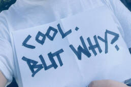 Person mit weißem T-Shirt hält ein Schild mit der Aufschrift „Cool. But Why?“ in schwarzer Schrift.