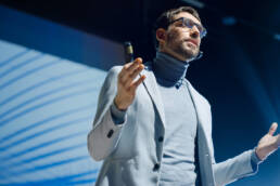 Mann in Rollkragenpullover und Brille hält Vortrag mit Mikrofon und Fernbedienung vor blau beleuchtetem Hintergrund.
