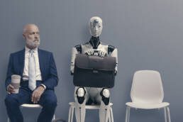Mann im Anzug mit Kaffeebecher sitzt neben Roboter mit Aktentasche auf Stühlen vor grauer Wand.